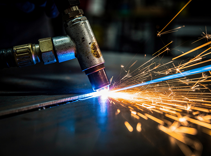 Macro action photograph of a plasma cutter torch slicing through 1/4 inch steel plate with bright blue arc and orange sparks flying with a clean narrow kerf line behind the torch