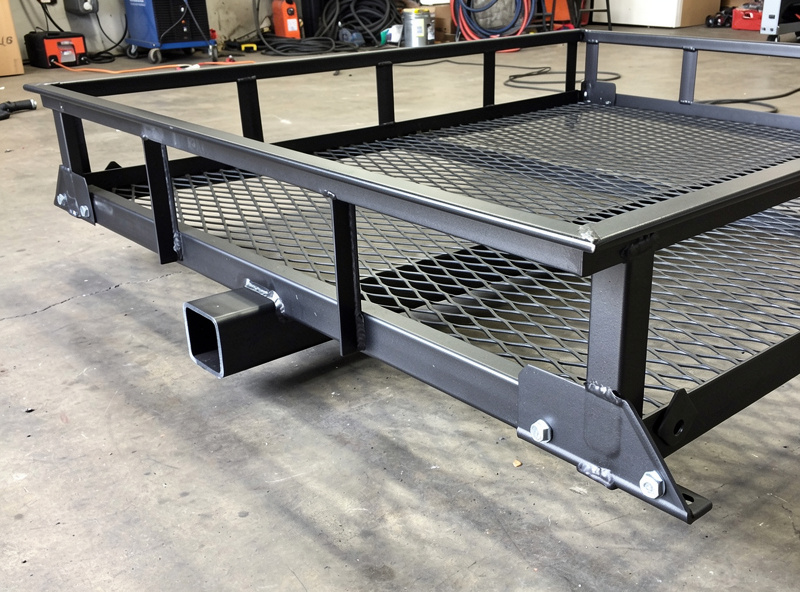 Photograph of a completed steel hitch-mounted cargo carrier on a workshop floor showing the receiver tube, platform frame with expanded metal deck, and corner gusset plates