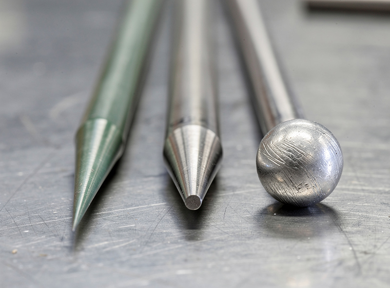 Macro close-up comparison of three tungsten electrodes on a workshop bench: leftmost sharp needle-point for DC TIG, middle truncated cone for AC TIG aluminum, right rounded ball tip showing improper grinding