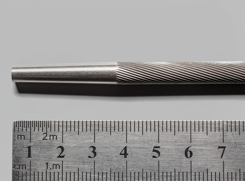 Side profile of a sharpened tungsten electrode showing 2.5 times diameter grind length with longitudinal scratch pattern parallel to the electrode axis under directional light with ruler for scale