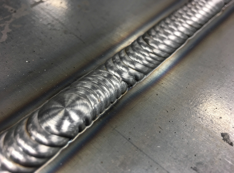 Macro photograph of a perfect MIG weld bead on mild steel showing consistent stacked-dime ripple pattern with no spatter on adjacent metal