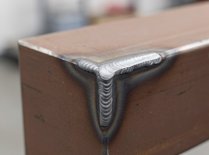 Two square steel tubes meeting at a tee joint with a freshly laid MIG weld bead, slight ripple pattern