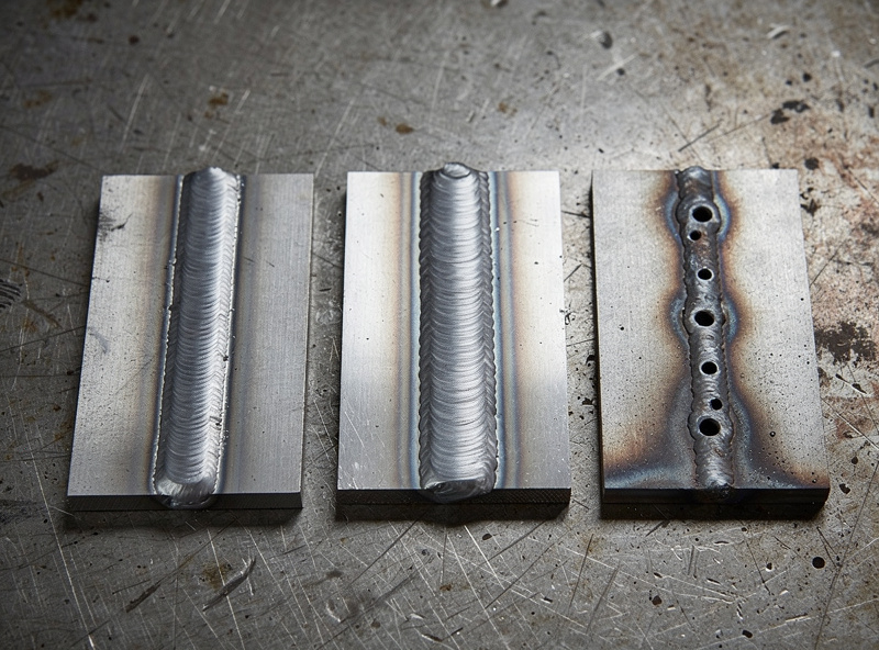 Three steel test coupons on a workbench showing different MIG weld bead profiles: cold weld with poor penetration, correct settings with even profile, hot with burn-through