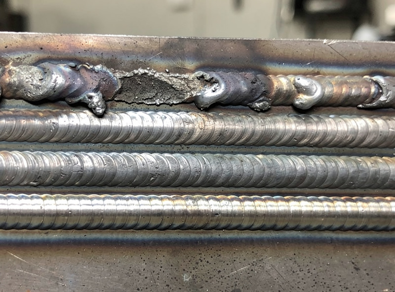 Close-up photograph of practice weld beads on a flat steel plate showing progression from beginner wobbly beads at the top to increasingly straight and consistent beads at the bottom, workshop setting