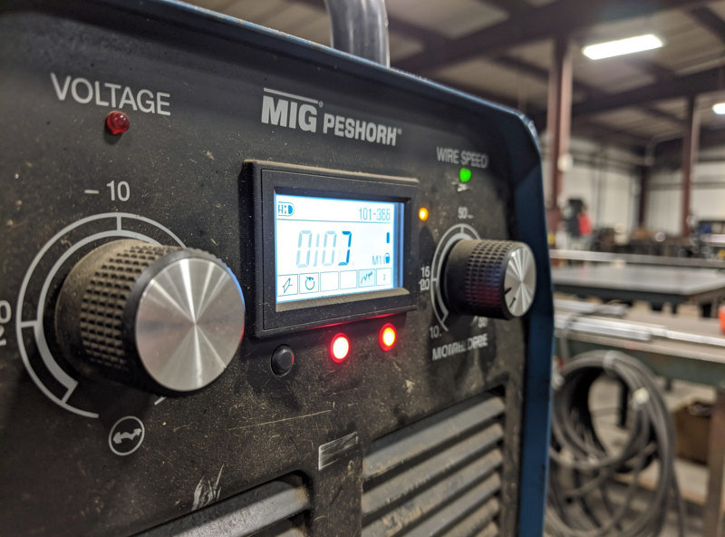 Close-up photograph of a MIG welder front control panel showing voltage knob, wire speed dial, and digital display with settings visible