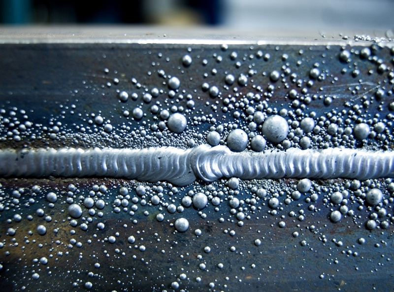 Excessive spatter balls on steel surface around a MIG weld bead showing clean bead contrast