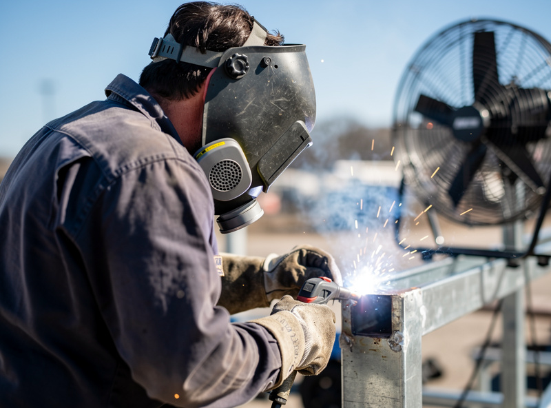 Welder welding galvanized steel outdoors with a fan and respirator, fume blowing away