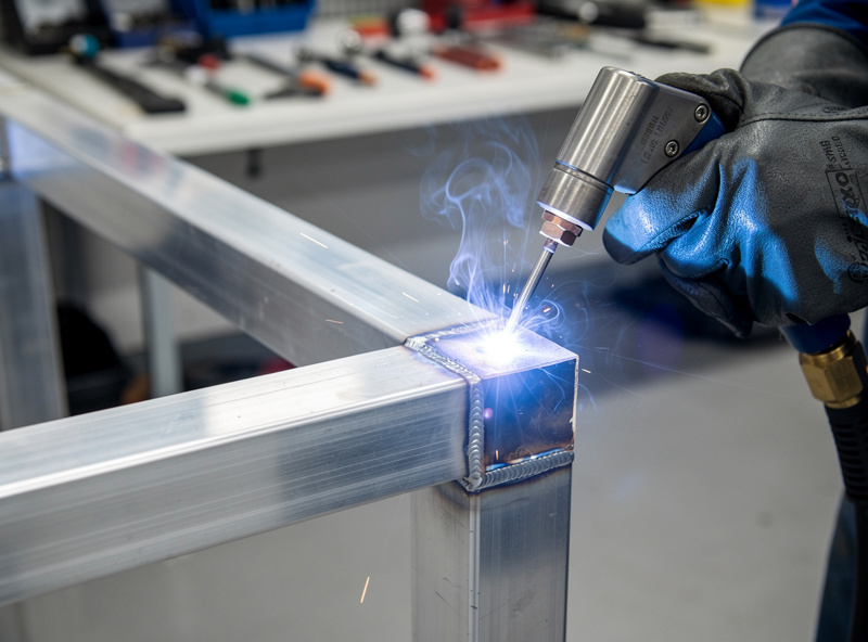 Aluminum welding on square tube frame with spool gun, argon shielding gas visible, clean workshop, metal fabrication detail