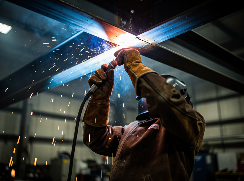 Welder MIG welding an overhead steel joint with sparks falling downward wearing full leather protection
