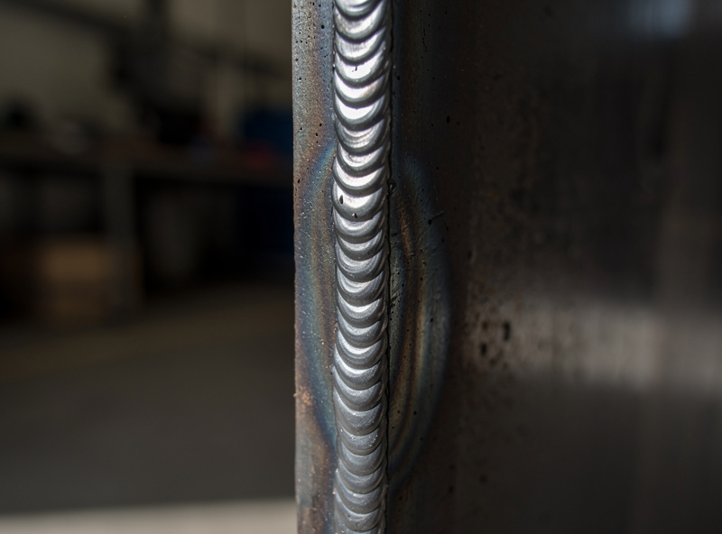 Close-up of a small controlled short-circuit MIG puddle on a vertical steel joint showing tight bead