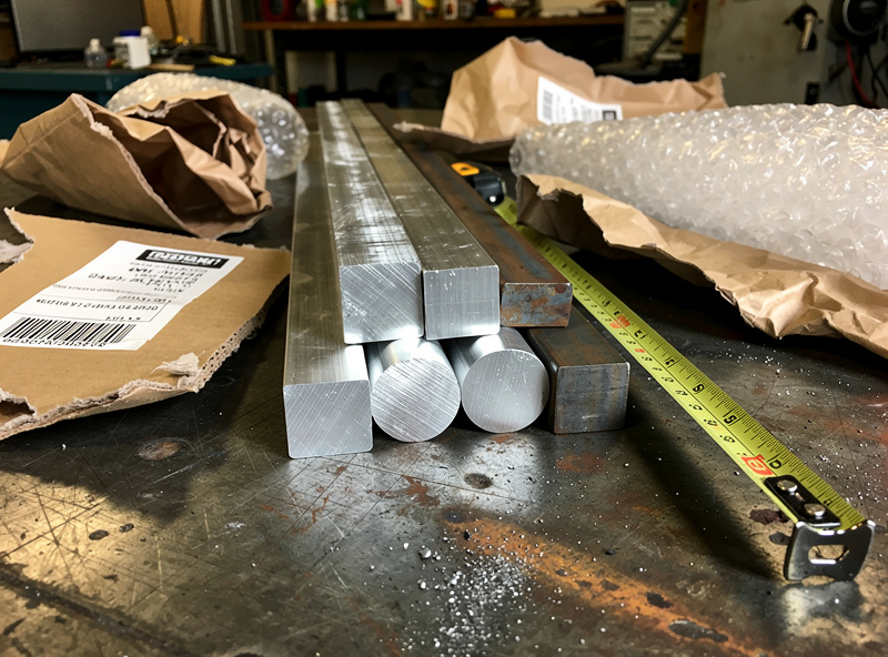 Cut-to-size aluminum and steel bar stock pieces freshly unboxed on a welding bench with shipping labels and packing material