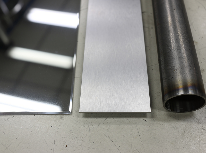 Three stainless steel samples on a welding bench - polished 304 sheet, 316 marine plate, and dull 409 exhaust tubing - with stainless filler rods