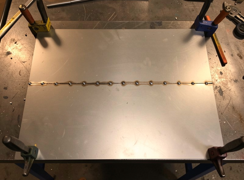 Thin sheet metal panel clamped flat to a welding table with a row of evenly spaced tack welds
