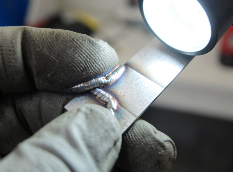 Gloved hand inspecting the bead profile of a welded steel test coupon under a work light