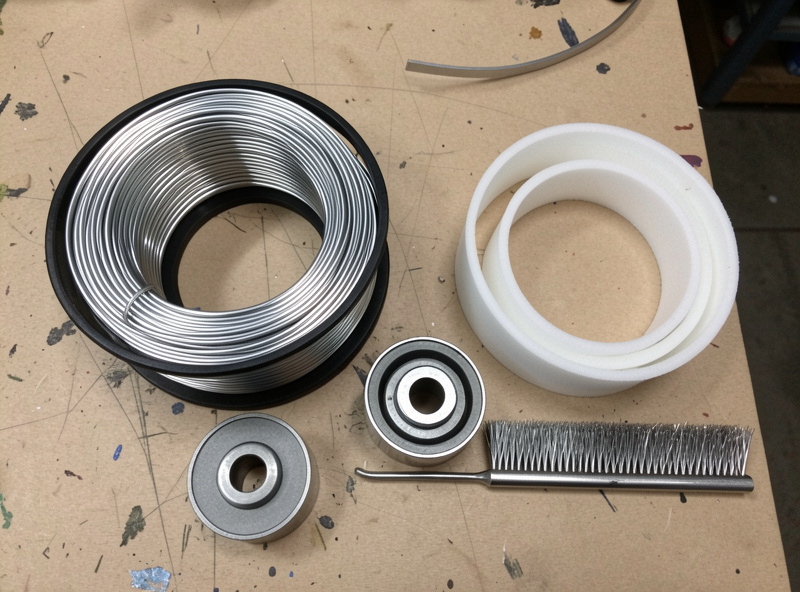 Flat-lay comparison of ER4043 and ER5356 spools, U-groove rollers, PTFE liner, and a stainless brush