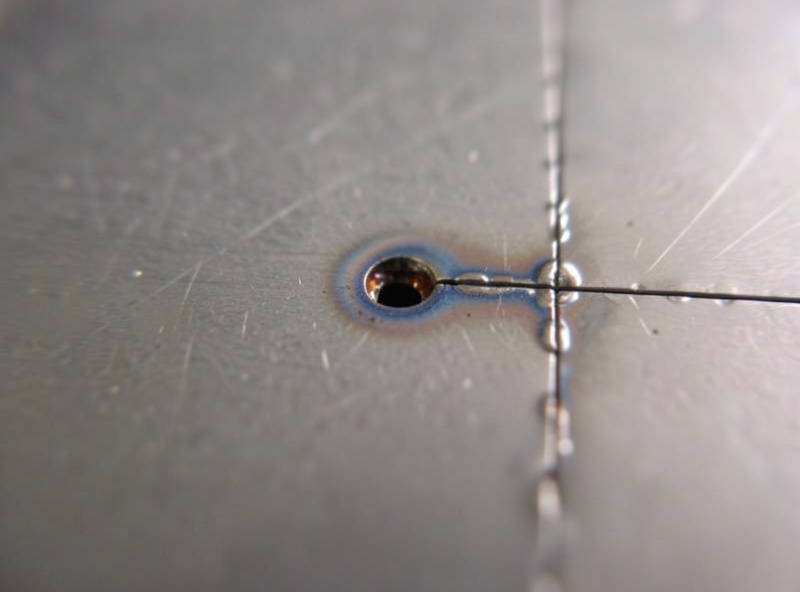 Thin steel sheet with a flux-core burn-through hole next to a successful stitch weld